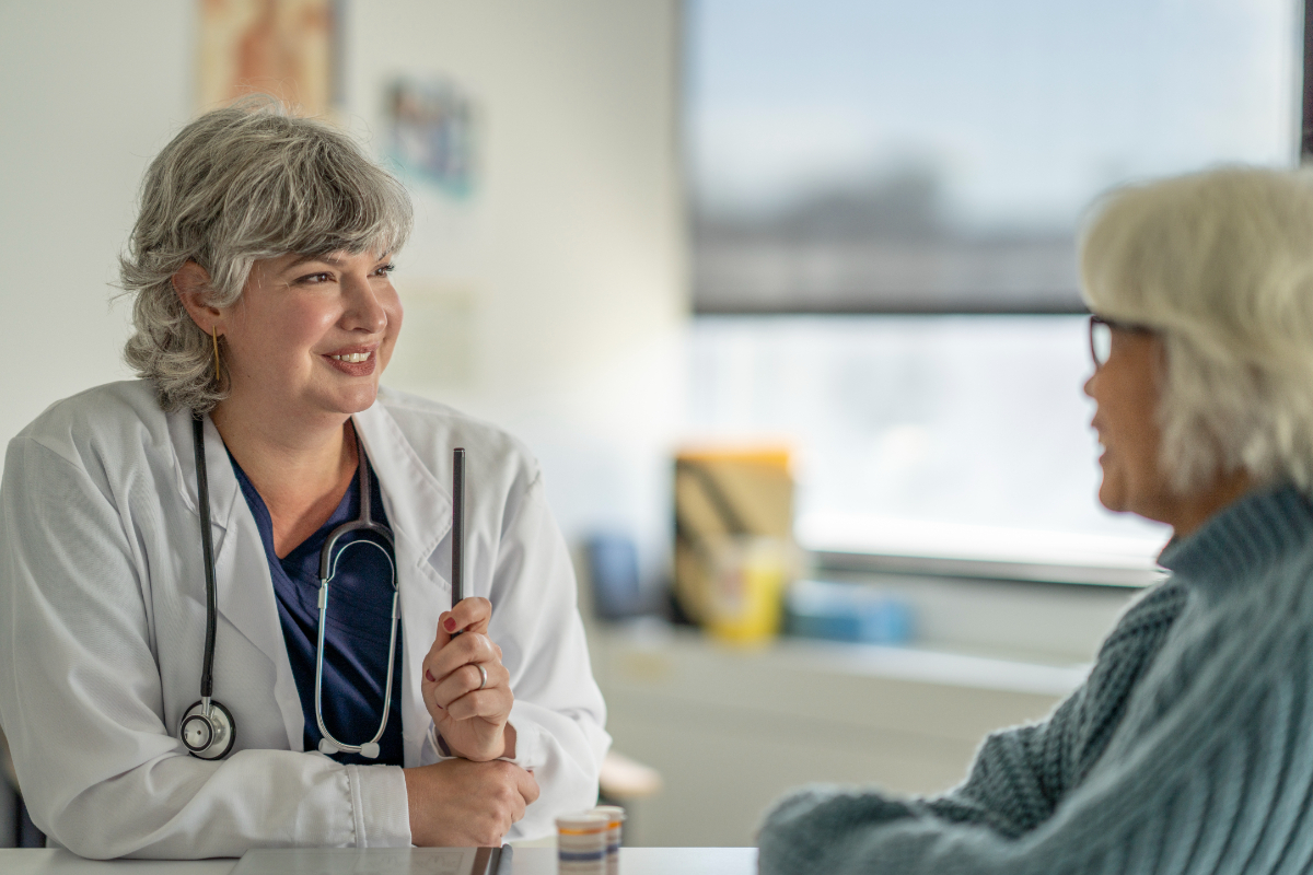 A female doctor sits across her desk from a senior patient as she discusses her recent test results. She is wearing a white lab coat and has a tablet out on the desk in front of the two as they discuss the results.